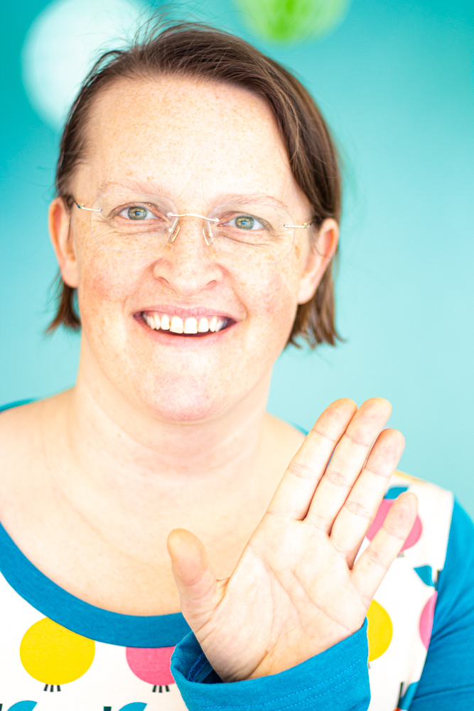 Stefanie Wolf with a self-made shirt with bold retro pink, yellow and orange apples on a vintage beige background. Stefanie is waving towards the viewer and smiling friendly.