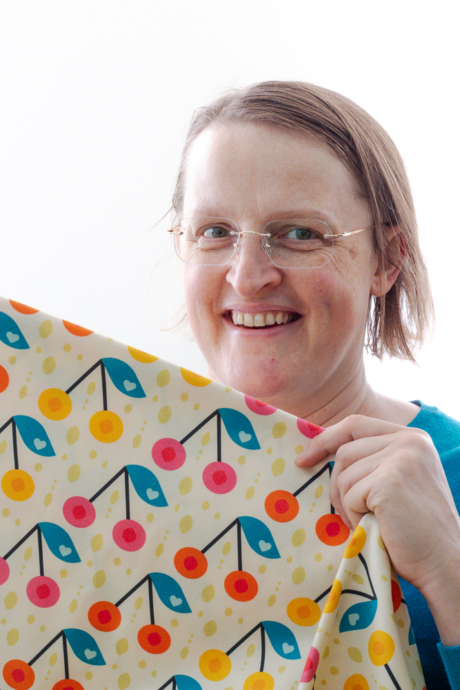 Stefanie Wolf smiling and holding a piece of self designed fabric in front of her. The fabric has bold retro orange, soft pink and warm happy yellow cherries in pairs with blue foliage with hearts on it.