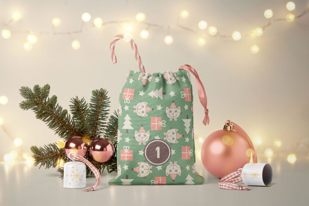 Fabric Advent calendar bag with beige white cats with party hats on a sage green background with peach pink gifts, Christmas trees, xmas stars. The bag is decorated with a peach pink beige white candy cane, a fir branch with 2 little pech pink Christmas baubles, a big Christmas tree ball and two littles bells on a beige white background illuminated with a chain of lights.