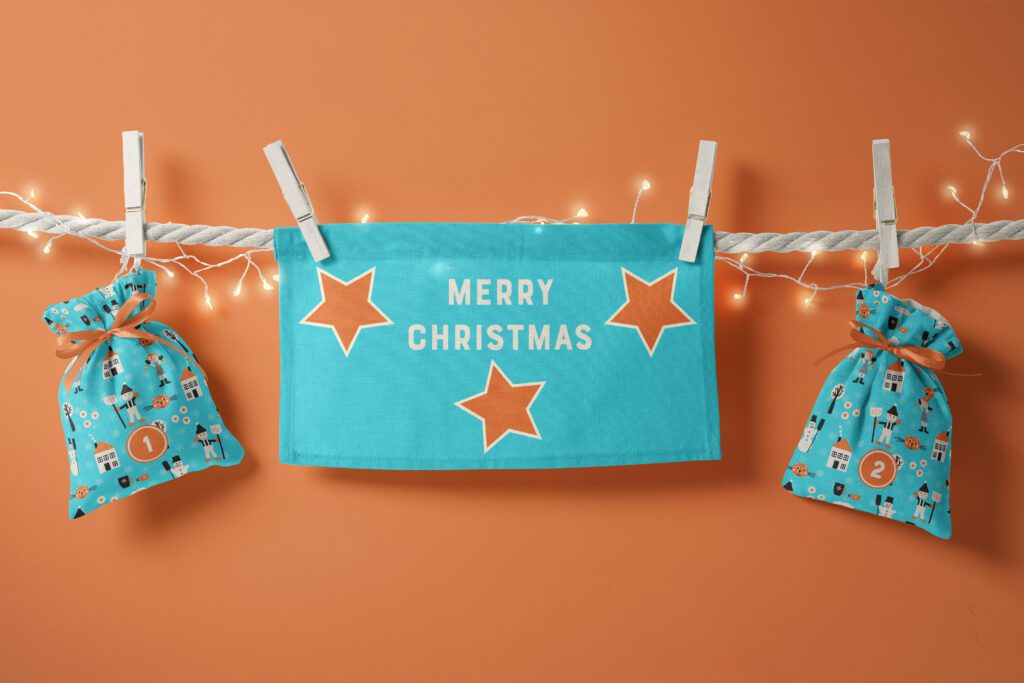 Two fabric Advent calendar bags in aqua blue with boys, girls, houses, cats, cookies, trees and snowflakes in orange, black and white hanging down froma beige white string on a terracotta orange wall. Everything is decorated with a Christmas-y chain of lights and a aqua blue little fabric flag with 3 orange stars and "Merry Christmas" on it.