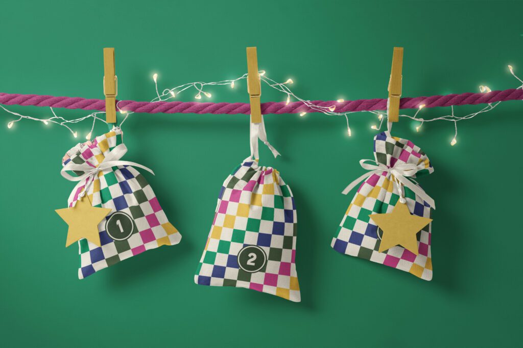 Three filled fabric Advent calendar bags with a bold, bright checkerboard pattern in hot feminine pink, mustard yellow, sage, jungle green and vintag beige white hanging down from a purple pink string decorated with a Christmas-y string of lights on a dark forest green wall.
