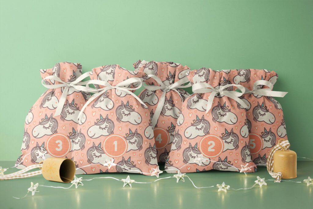 Five filled Advent calendar bags with cute hand drawn unicorns on a kitschy pastel 1950s background with little hearts. The bags are closed with little cream white ribbons and stand on a sage green background decorated wit a Christmas-y string light and little bells.