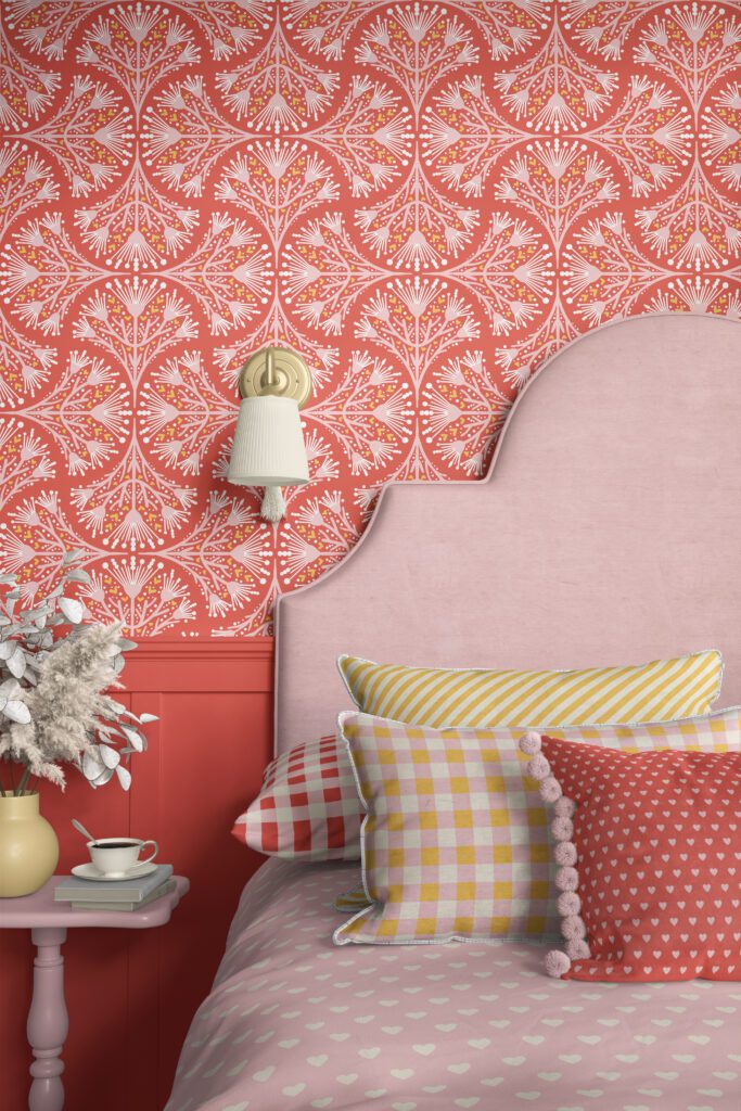 Romantic feminine bedroom with decorated with patterns from the "Vintage Daydream" collection. As soft pink bed with a velvet headboard, a pink duvet cover with little white hearts, and several pillows with pink, yellow, red and beige gingham checks. A red pillowe with little soft pink hearts. And a pillow with diagonal beige white and warm yellow stripes. The wall is decorated with a coral red wallpaper with geometric soft pink, beige white and yellow flowers in a non-directional pattern.