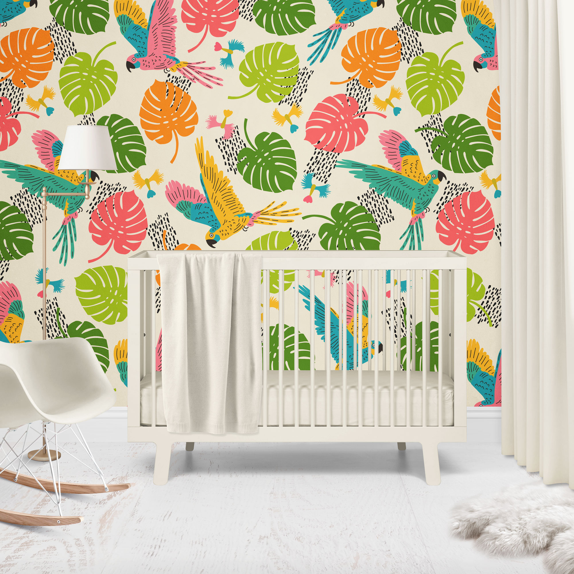 Baby-nursery---joyful-jungle---flying-parrots-in-colourful-monstera-leaves-M Nursery wallpaper in a playful, colorful tropical style featuring flying parrots and vibrant monstera leaves in pink, green, orange, yellow, and blue on a cream background. The scene includes a white baby crib with a draped blanket, a modern white rocking chair, and a floor lamp, creating a bright and cheerful nursery setting.