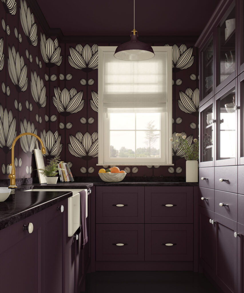Dark plum kitchen with matching aubergine cabinets and ceiling, featuring a traditional non-pasted wallpaper in deep aubergine with large-scale beige graphic florals, black countertops, brass faucet, and a window with a white Roman shade above a bowl of fruit and a tall white vase of greenery.