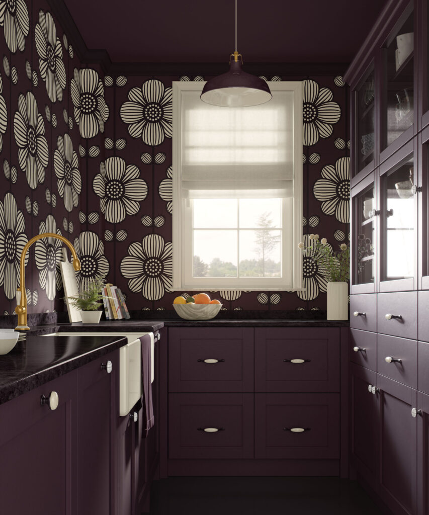 Bold dark plum kitchen with traditional non-pasted wallpaper on the walls and ceiling, featuring large beige graphic flowers, matching aubergine cabinets with black countertops, a farmhouse sink, brass faucet, white roman shade over the window, and a bowl of fruit on the counter.