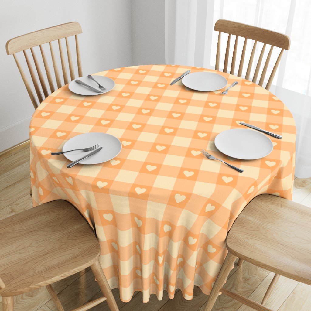 A tablecloth with a gingham check pattern in warm cream and soft apricot orange colors, featuring subtle heart shapes within the checks. The tablecloth is draped over a round wooden dining table surrounded by light wood chairs, set in a bright, airy kitchen or dining space with natural light and minimal decor.