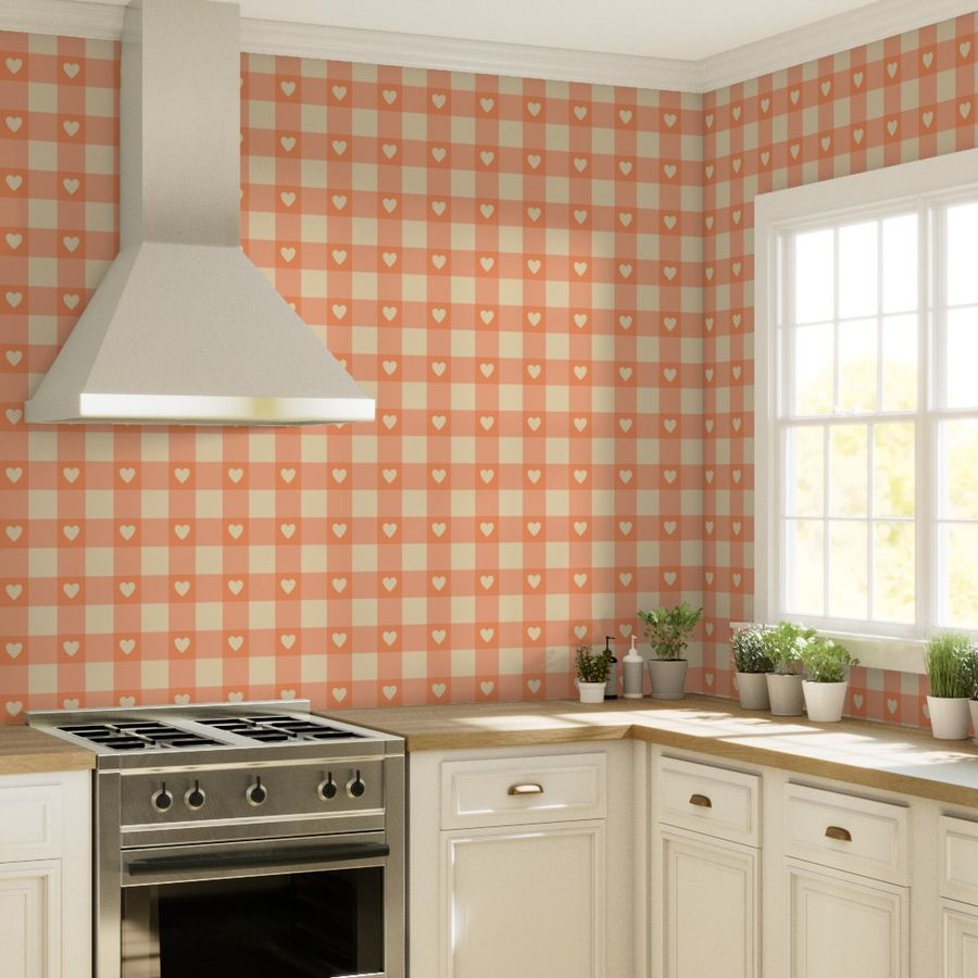 Wallpaper featuring a warm tangerine and beige gingham check pattern with small heart shapes in a bright kitchen. The kitchen includes white cabinetry, a stainless steel stove, wooden countertops, and several potted plants by a large window letting in natural light.